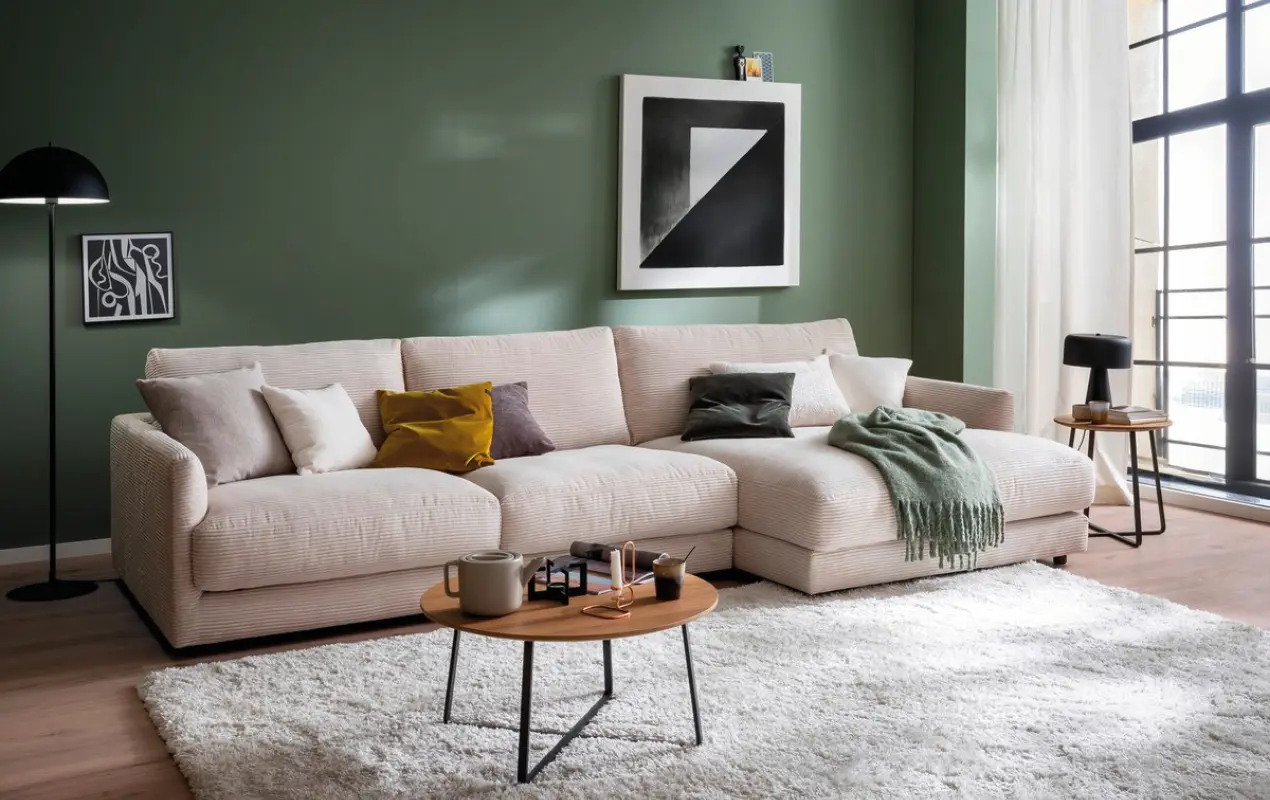 Helles Ecksofa mit Kissen, Couchtisch und Teppich vor grüner Wand und Fenster